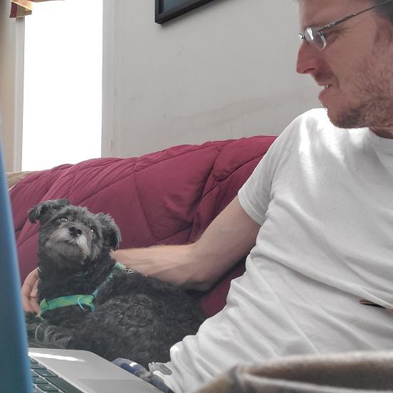 Jimmy sits on his sofa with a laptop on his lap. He has one arm on the small dog by his side, his Poodle Pomeranian named Rudy. Rudy and Jimmy look at each other fondly.
