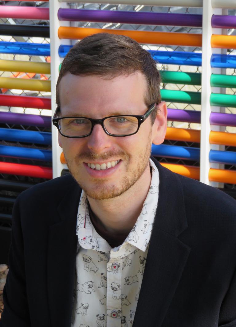A headshot of Jimmy Matejek-Morris. Image taken in front of a fence made of horizontal rainbow pipes.